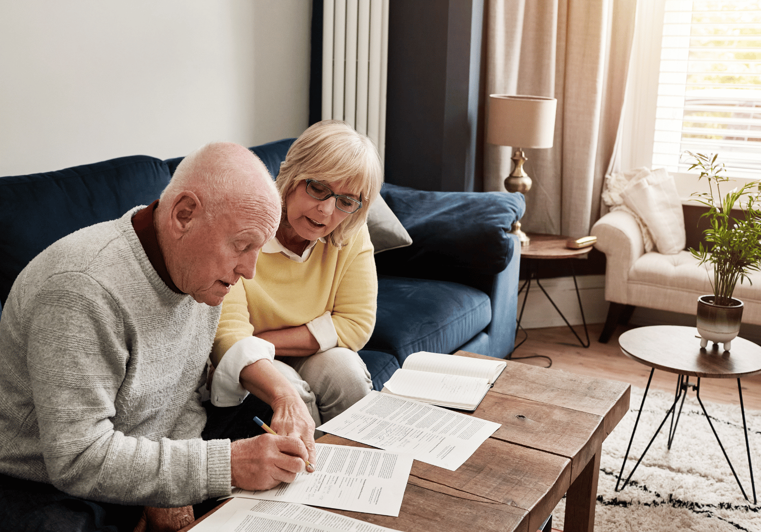 Elderly couple sitting on a blue sofa, reviewing documents spread on a wooden coffee table.