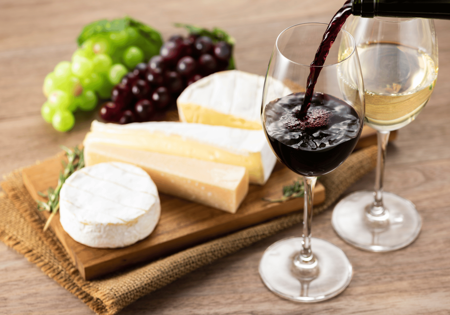 Red wine is poured into a glass beside a cheese board with brie and other cheeses and grapes on a wooden table.